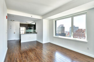 Unit living room, hardwood floors, windows on the right in photo, photo taken in living room towards kitchen and entryway door, white walls, windows with view of the city.