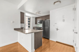Unit kitchen, stainless steel appliances, hardwood floors, window in kitchen, green subway tile between cabinetry and countertop, entry door adjacent to kitchen, breakfast nook attached to kitchen countertop, dark brown wooden cabinetry, white walls.