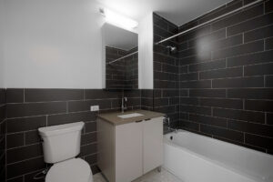 Unit bathroom, bathtub/shower, vanity with storage, sink with storage underneath, tile floor, dark colored subway tile on walls.