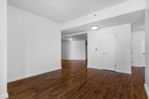 Unit Living room, open floorplan, hardwood floors, storage closet adjacent to living room and kitchen, entry walkway leading to living room then kitchen, White walls, overhead lighting.