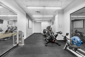Community Fitness room, white walls, gray matted carpet, elliptical machines, stationary bicycles, rowing machine, TVs above elliptical machines, stretching area.