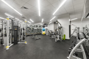Community Fitness room alternate view, white walls, gray matted carpet, weightlifting section, free weight section, multiple machines.