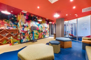 Community Kids Room, multicolored walls, salmon colored ceiling, children’s playset, children’s Basketball hoop, Hexagonal furniture, misc. toys, waiting area outside for parents to watch.