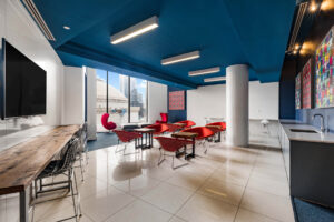 Community Lounge Room, 4-2 chair tables, bar seating along perimeter, kitchen sink, TV, Tile and Carpeted Floor, Square contemporary art on white & blue walls, floor to ceiling windows with view of city, seating near window.