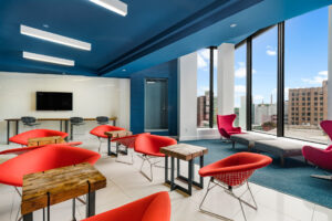 Community Lounge Room alternate view, 4-2 chair tables, bar seating along perimeter, kitchen sink, TV, Tile and Carpeted Floor, Square contemporary art on white & blue walls, floor to ceiling windows with view of city, seating near window.