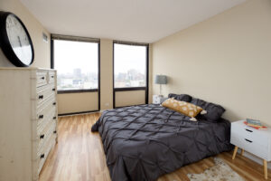 Unit Bedroom, Furnished, queen size bed, 2 nightstands, wood floors, large dresser, 2 large windows with view of downtown.