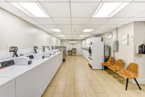 Interior Laundry Facility, rows of industrial washing machines and dryers, tile floor, three seat bench, industrial sink, white walls.