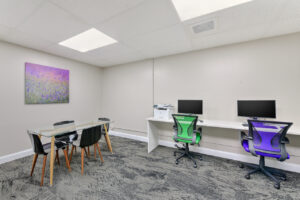 Interior business center four computer stations, printer, glass table with four seats, contemporary carpeted floor, white walls, floral painting on the wall.
